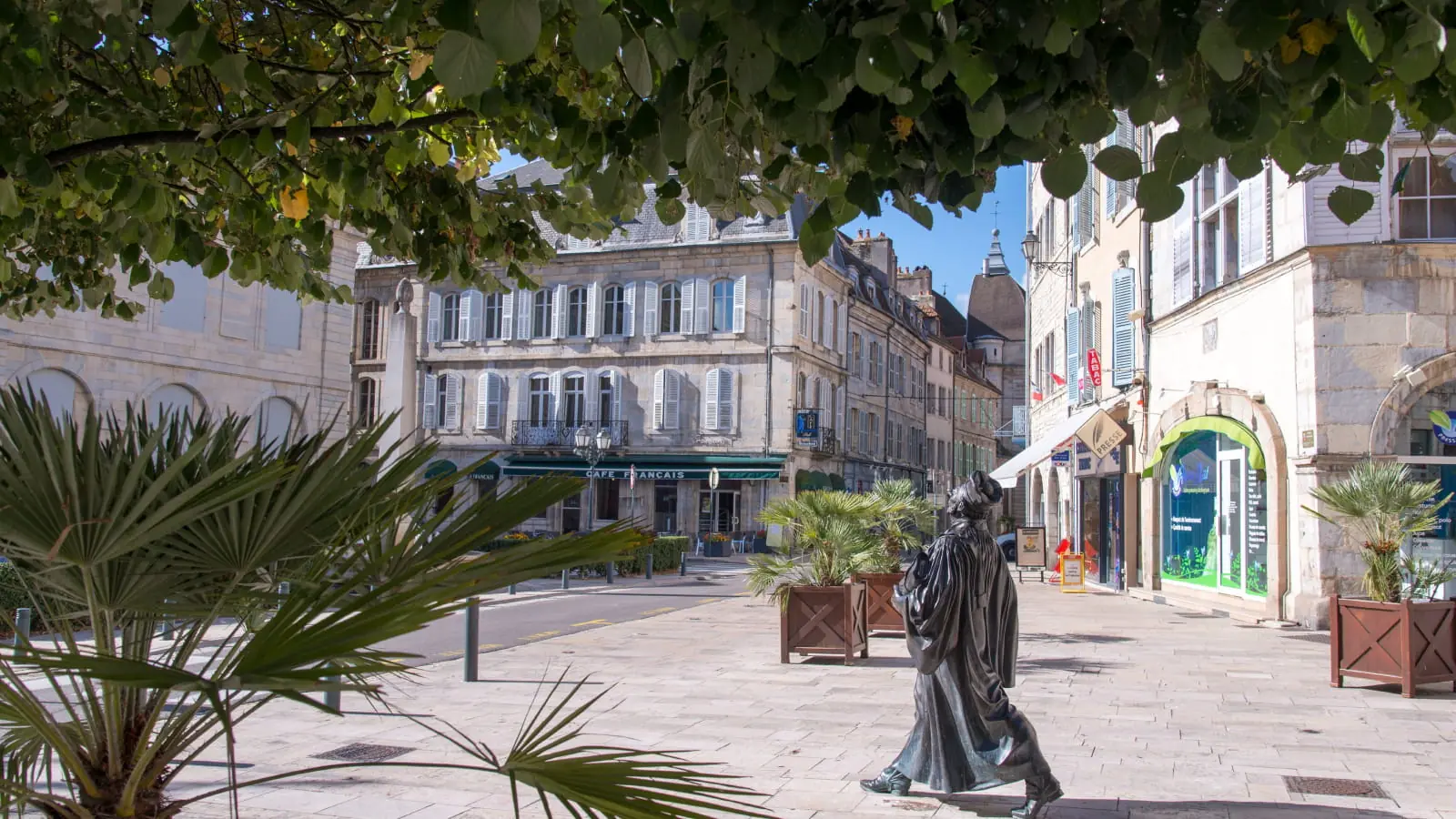 Le centre ancien de Vesoul : façades de pierre à volets clairs, terrasse de café et statue de bronze sur une place pavée.